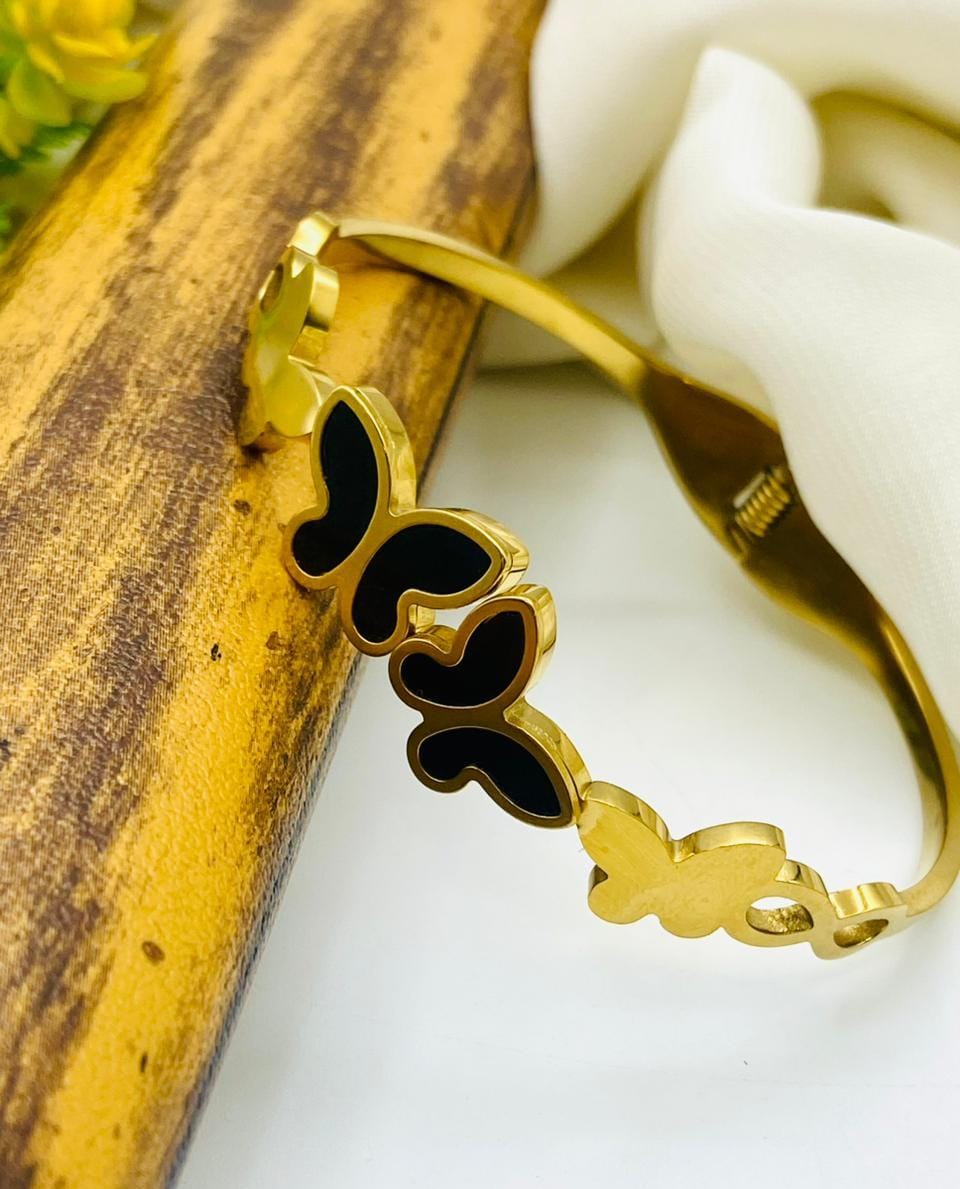 A gold-plated bangle bracelet featuring a continuous butterfly motif with black enamel inlay, resting on a rustic wooden block and a white fabric background.