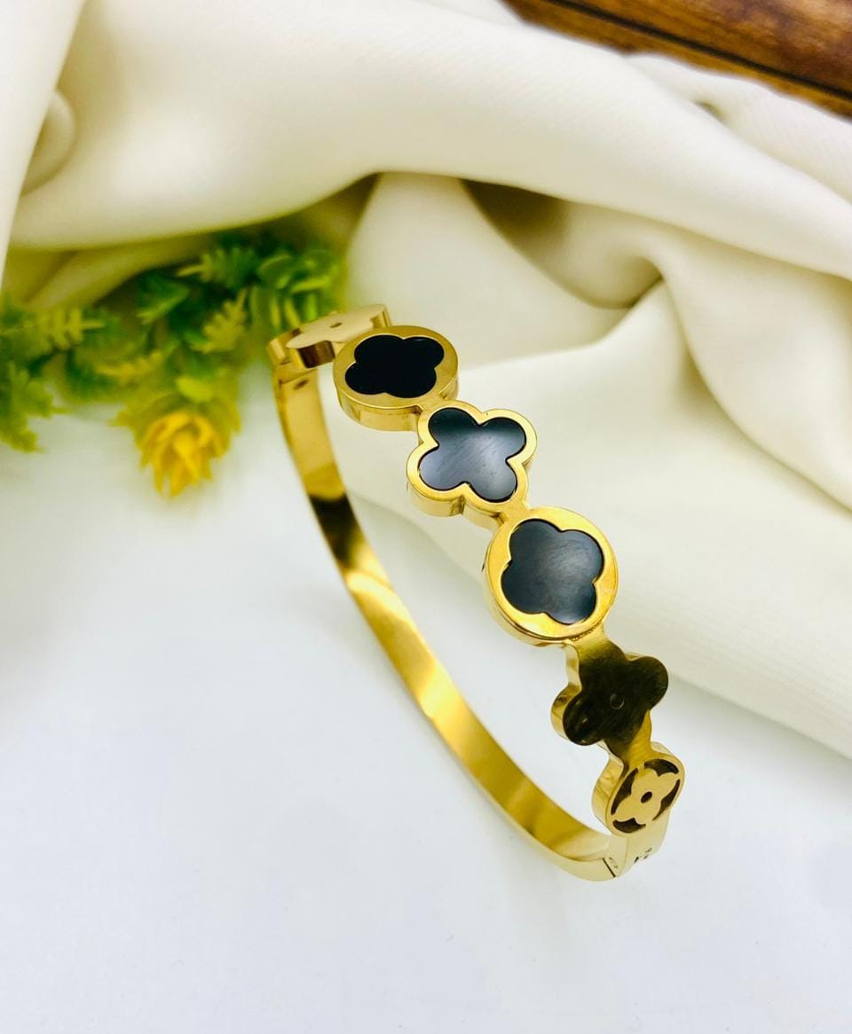 A gold-plated bangle bracelet featuring four clover-shaped motifs with black inlay, styled on a white background with draped cream fabric and small green leaves.