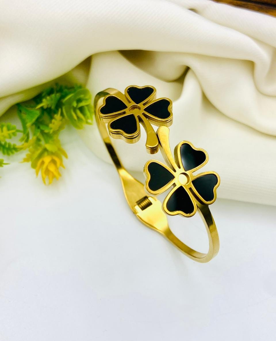 Gold-tone cuff bracelet with two four-leaf clover motifs filled with black enamel, styled on a white surface with cream fabric and small green leaves.