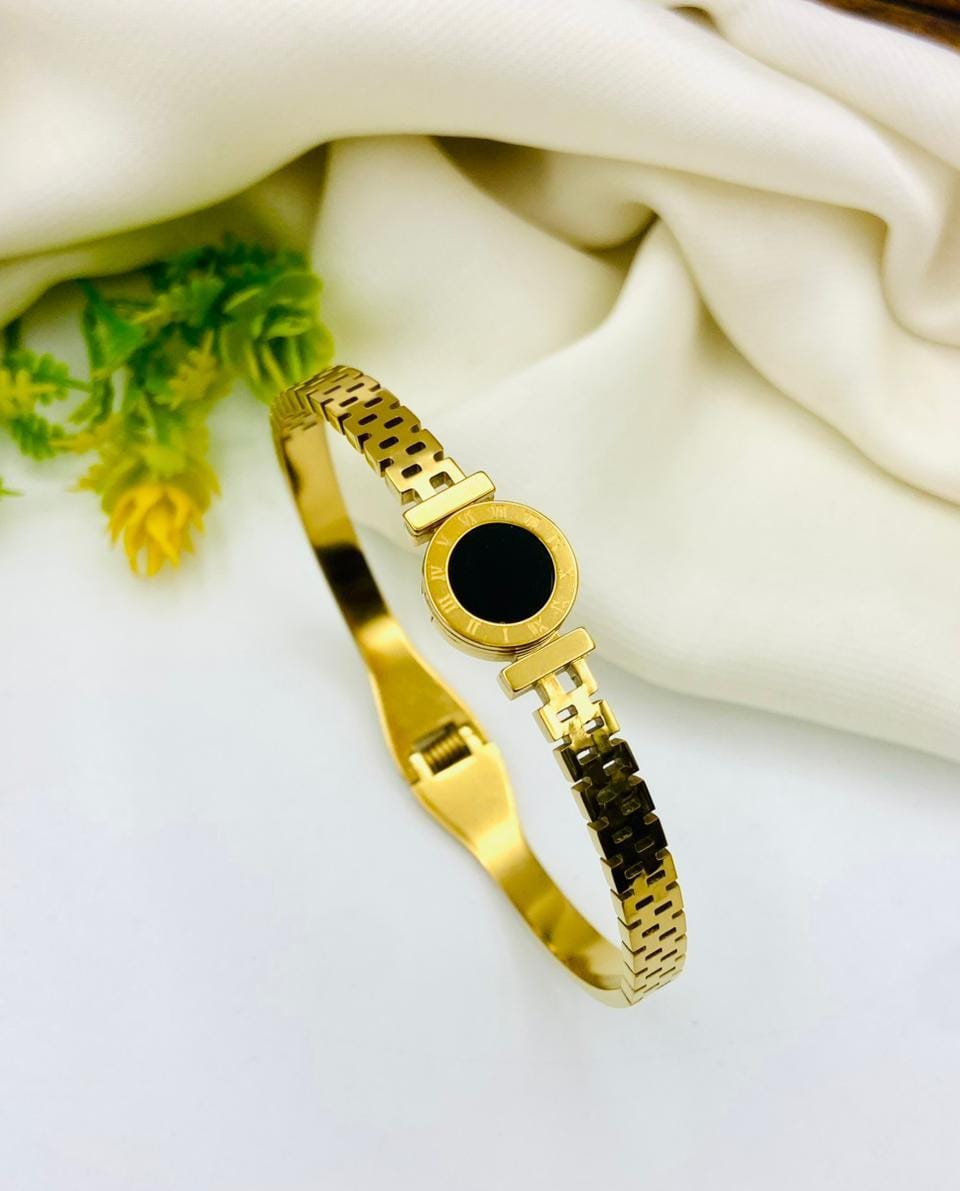 A gold-plated metal bangle watch with a round black dial, styled on a white surface with draped cream fabric and a small green plant in the corner.
