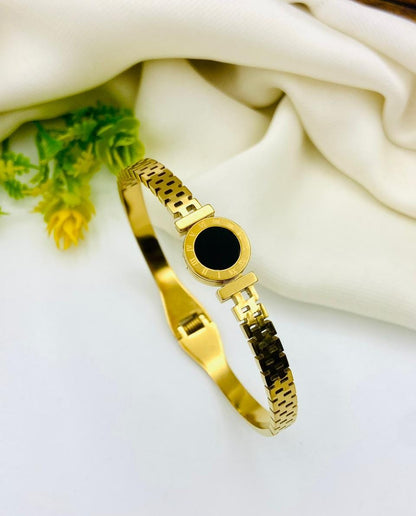 A gold-plated metal bangle watch with a round black dial, styled on a white surface with draped cream fabric and a small green plant in the corner.