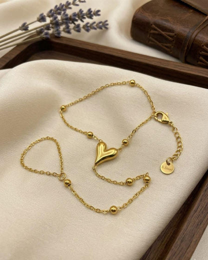 A delicate gold chain bracelet with a polished heart charm, beaded accents, and an adjustable sliding clasp, displayed on a cream fabric surface with a wooden tray and dried lavender in the background.