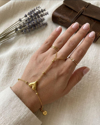 A gold hand chain bracelet with a heart-shaped charm and a small dangling disc, shown worn on a person's hand alongside gold rings, set against a neutral linen background with dried lavender and a brown leather journal.