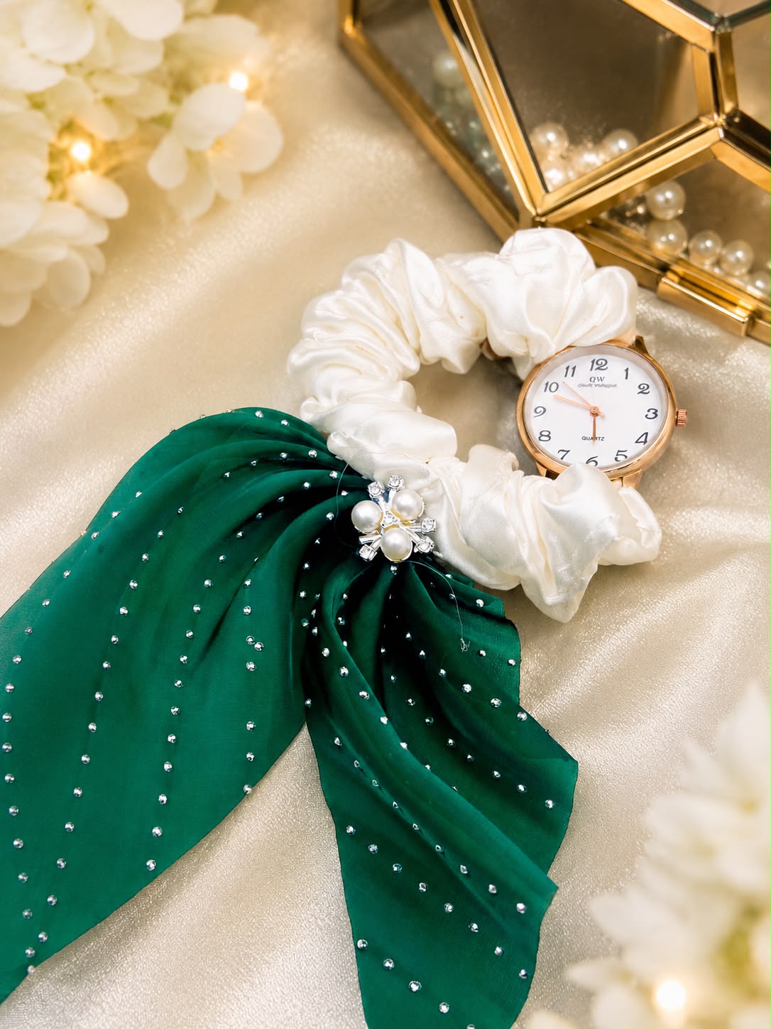 A wristwatch with a rose gold case and white dial, styled with a white fabric scrunchie and a green rhinestone-studded scarf on a beige satin background.
