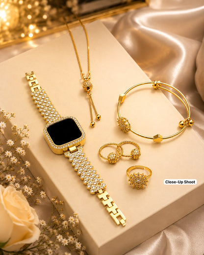 A gold-toned jewelry set arranged on a cream display block, including a smartwatch-style bracelet watch with a rhinestone-embellished link band, a delicate necklace, a bangle bracelet, two rings, and a cluster of small stud earrings. The set is styled with satin fabric, blurred lights, and small flowers.