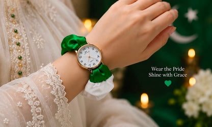 A close-up lifestyle shot of a woman's wrist wearing a watch with a white dial and a green satin ribbon bracelet. The person is wearing a white lace-trimmed dress, with a festive green and white background featuring warm bokeh lights and the text 'Wear the Pride Shine with Grace'.