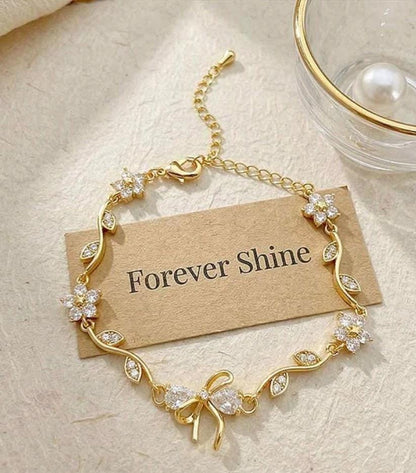 A gold chain bracelet with floral and bow motifs set with clear rhinestones, displayed on a beige card that reads 'Forever Shine' on a textured cream surface.